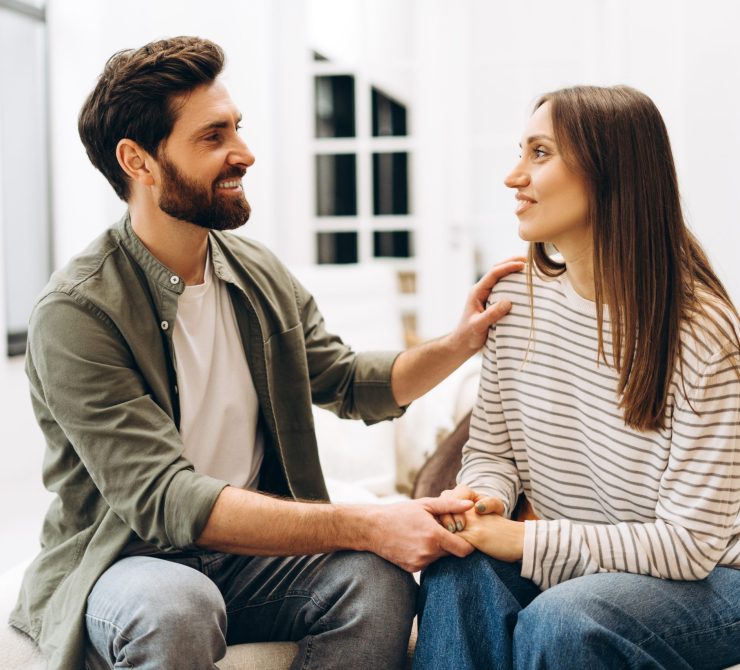 Male psychologist supporting female patient by holding hands and comforting her during therapy session on the sofa, showing empathy and care